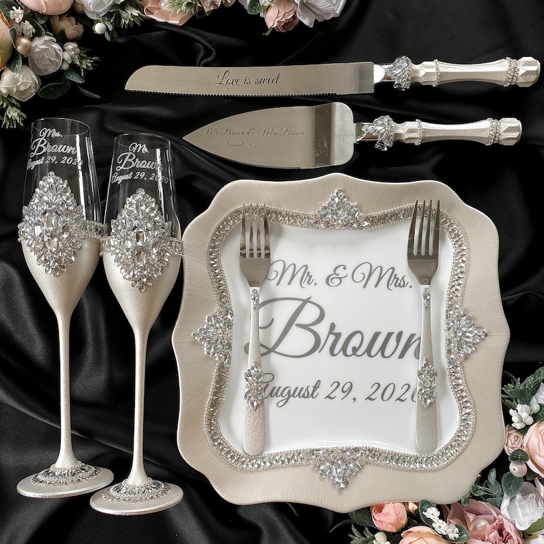 Puede incluir: Un juego de servir pastel de bodas con un plato de pastel blanco, dos tenedores y un cuchillo. El plato tiene un borde decorativo de diamantes de imitaci&oacute;n y el texto "Mr. & Mrs. Brown August 29, 2020". Tambi&eacute;n se incluyen dos copas de champ&aacute;n con diamantes de imitaci&oacute;n. El cuchillo y la esp&aacute;tula de servir tienen mangos plateados con diamantes de imitaci&oacute;n.