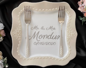 May include: White ceramic cake plate with a scalloped edge and pearl trim. The plate has the text "Mr. & Mrs. Mondur 08/12/2020" printed on it. Two silver forks are placed on the plate.