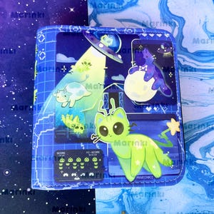 May include: A square blue wallet featuring a space-themed graphic print. The design includes a bright green alien cat, a cow being abducted by a UFO, a purple cat sitting on a moon, and a retro pixelated video game score display in the corner.