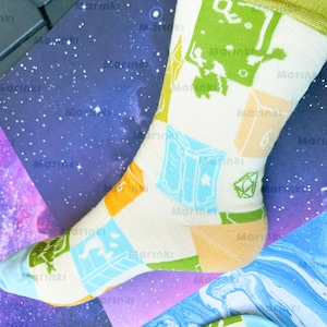 Peut inclure: Une chaussette de couleur crème avec un motif répétitif d'icônes sur le thème du jeu, incluant des livres bleu clair, des clés vertes et des dés orange avec le chiffre six imprimé. La chaussette possède un revers vert clair et une pointe bleue.