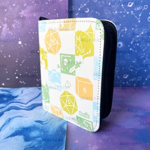 May include: A white zippered binder featuring a repeating grid pattern of light green, yellow, and blue icons including polyhedral dice, a potion bottle, a skull, and a dragon. The word Marinki is printed in small text on the bottom right corner.