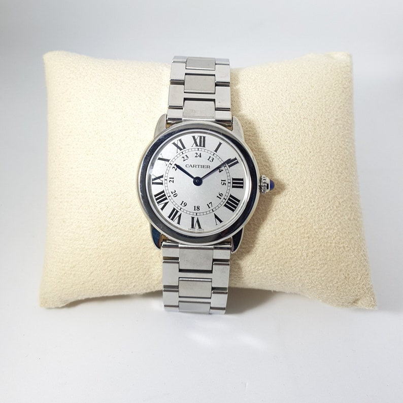 May include: A silver Cartier wristwatch with a white face and black Roman numeral hour markers. The watch has a blue sapphire cabochon on the crown. The watch is on a silver metal bracelet.