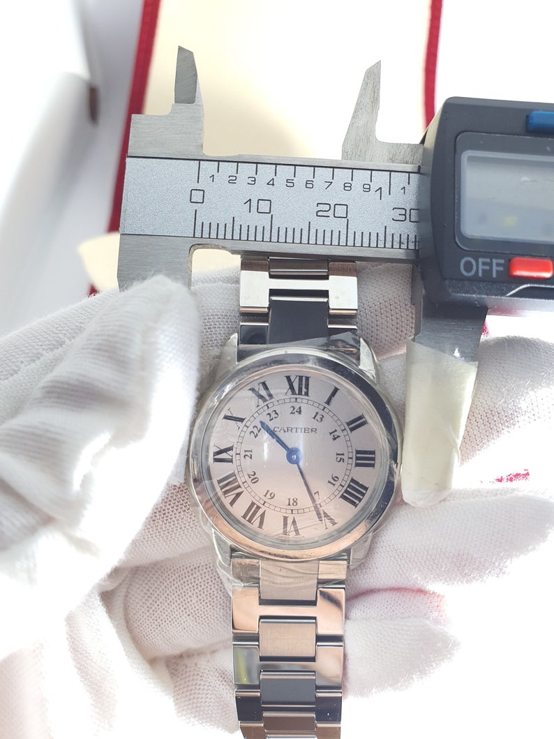 May include: A silver Cartier watch with a white face and black Roman numeral markings. The watch face also has black markings for the hours and minutes. The watch has a silver metal band.