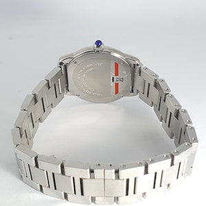 May include: A silver wristwatch with a blue sapphire on the crown. The watch has a silver band and a round, silver face with black markings. The back of the watch is visible and reads 'WATER RESISTANT 100M SWISS MADE STAINLESS STEEL'.