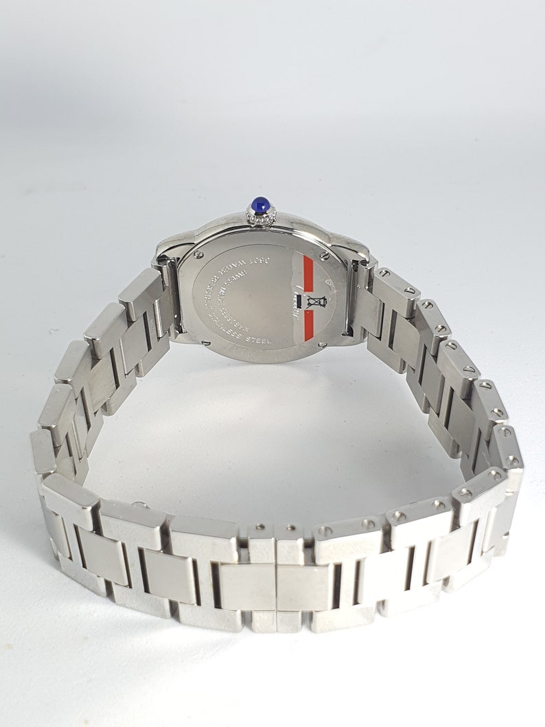 May include: A silver wristwatch with a blue sapphire on the crown. The watch has a silver band and a round, silver face with black markings. The back of the watch is visible and reads 'WATER RESISTANT 100M SWISS MADE STAINLESS STEEL'.