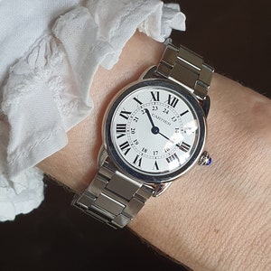 May include: A silver Cartier wristwatch with a white face and black Roman numeral hour markers. The watch has a silver metal band and a blue sapphire cabochon on the crown.