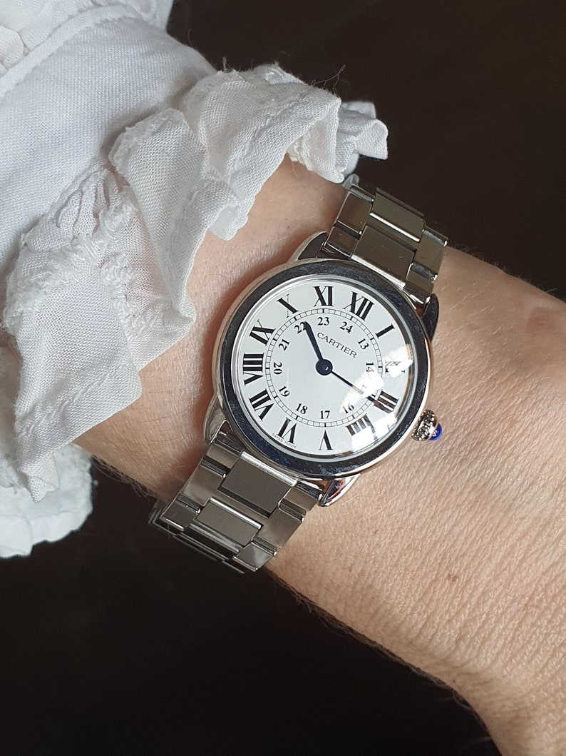 May include: A silver Cartier wristwatch with a white face and black Roman numeral hour markers. The watch has a silver metal band and a blue sapphire cabochon on the crown.