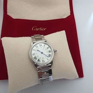 May include: A silver Cartier watch with a white face and black Roman numeral markings. The watch is in a white velvet watch box with a red velvet exterior. The exterior of the box has the word 'Cartier' printed in gold lettering.