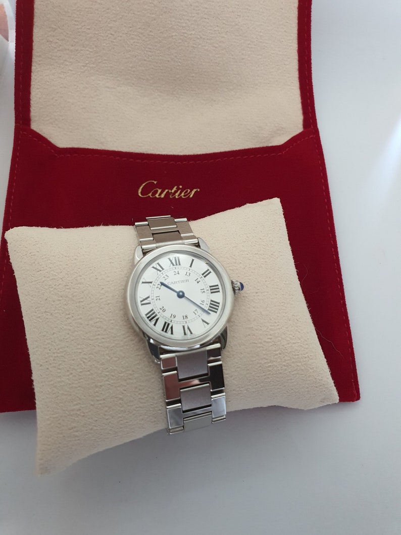 May include: A silver Cartier watch with a white face and black Roman numeral markings. The watch is in a white velvet watch box with a red velvet exterior. The exterior of the box has the word 'Cartier' printed in gold lettering.