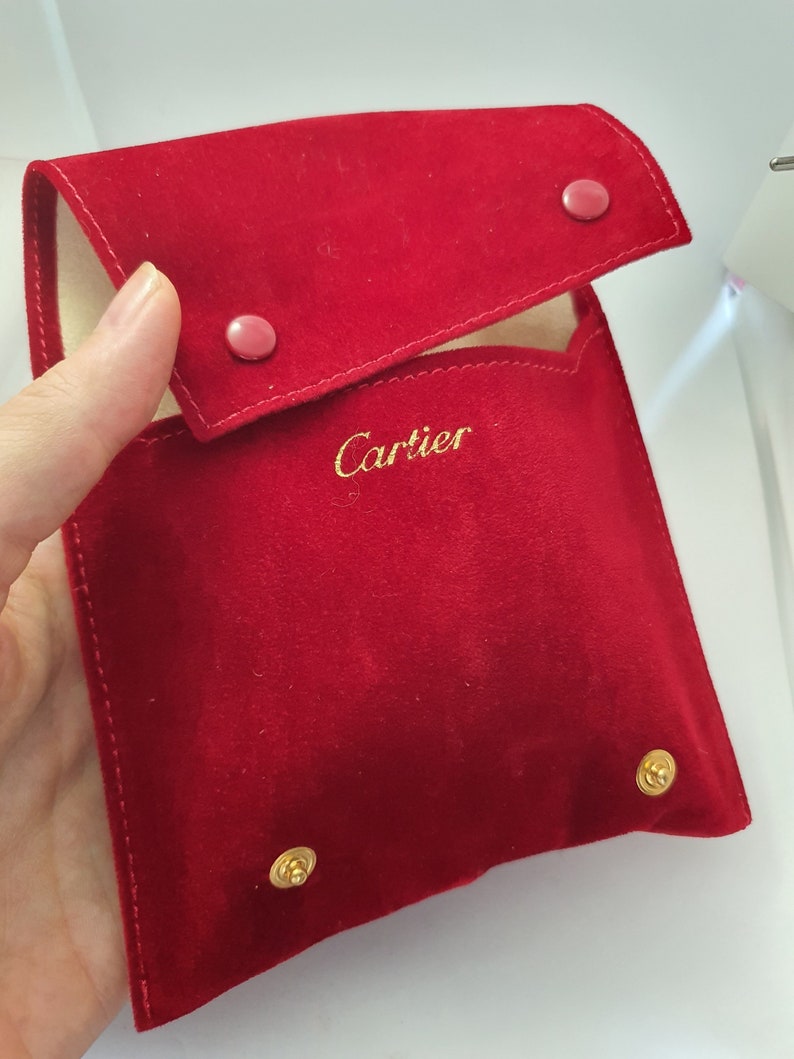 May include: A red velvet pouch with gold accents and the word 'Cartier' embroidered in gold on the front.