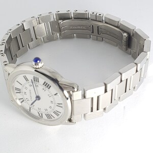 May include: A silver Cartier wristwatch with a white face and black Roman numeral hour markers. The watch has a silver metal band with a clasp that reads 'Stainless Steel'.
