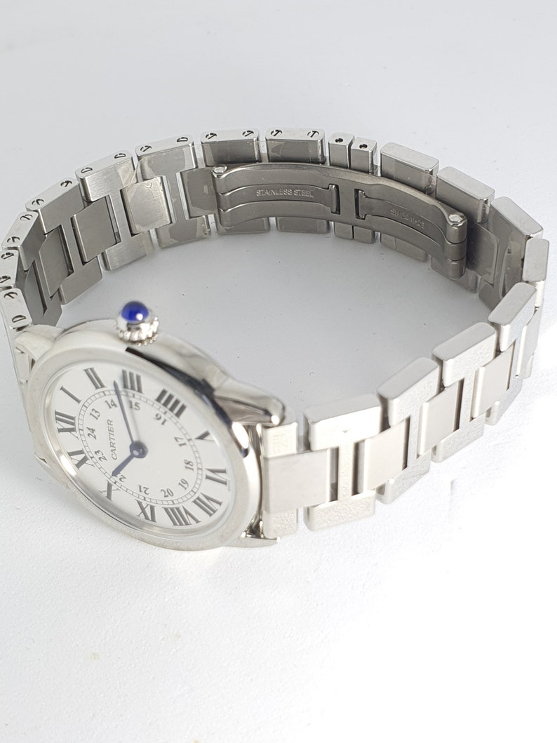 May include: A silver Cartier wristwatch with a white face and black Roman numeral hour markers. The watch has a silver metal band with a clasp that reads 'Stainless Steel'.