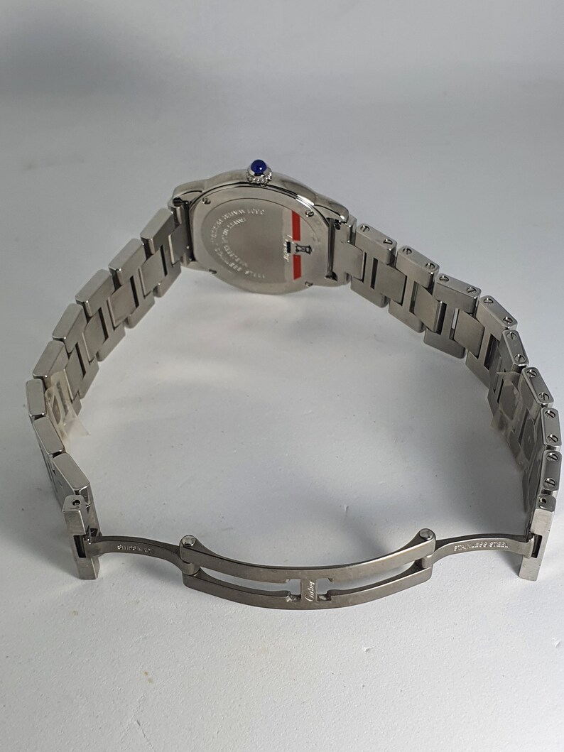 May include: A silver wristwatch with a silver band. The watch face is round and has a blue sapphire crystal. The back of the watch has the words 'Cartier' and 'Swiss Made' engraved on it. The watch band has a deployant clasp with the word 'Cartier' engraved on it.