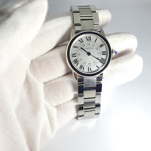 May include: A silver Cartier watch with a white face and black Roman numeral hour markers. The watch has a black outer ring with white numbers for the minutes. The watch face also has a black second hand and a black date window at the 3 o'clock position. The watch has a silver metal band.
