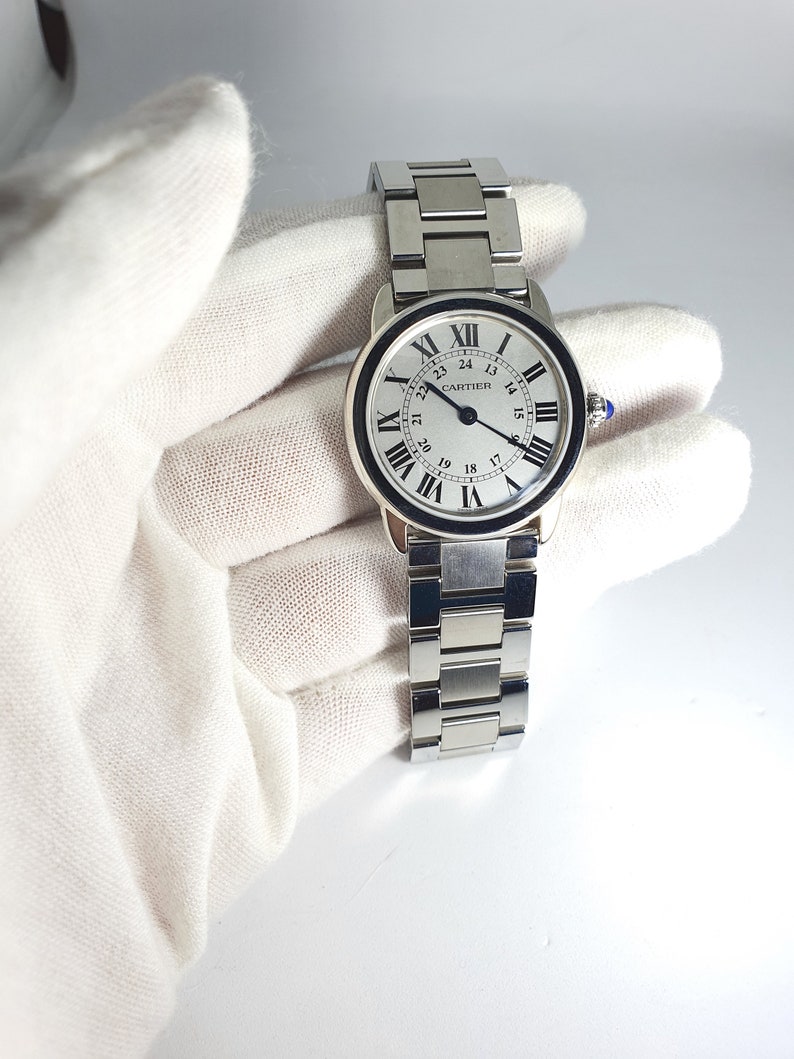May include: A silver Cartier watch with a white face and black Roman numeral hour markers. The watch has a black outer ring with white numbers for the minutes. The watch face also has a black second hand and a black date window at the 3 o'clock position. The watch has a silver metal band.