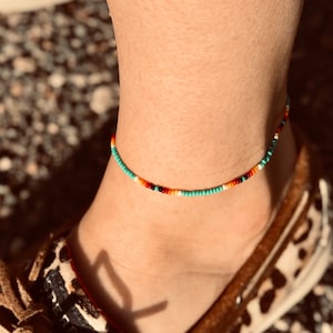 May include: A turquoise, orange, red, and black beaded ankle bracelet. The bracelet is thin and delicate, and it is worn on a person's ankle.