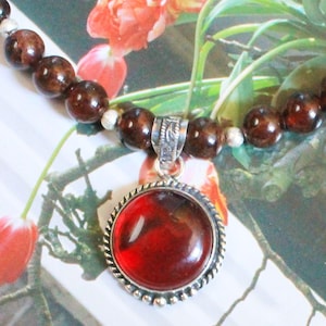 Peut inclure: Un collier avec un grand pendentif rond rouge bordé d'une bordure argentée en forme de corde. Le collier est composé de perles sphériques marron foncé et de petites perles d'espacement argentées. Le fond présente un motif floral.
