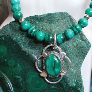 Peut inclure: Un collier avec un pendentif en argent orné d'une pierre ovale en malachite. Le collier est composé de perles rondes de malachite verte, avec des accents argentés. Le bijou est posé sur une surface en pierre verte.