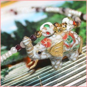 May include: A white enamel elephant charm with red, green, and gold floral details. The charm is attached to a beaded necklace with white and purple beads.