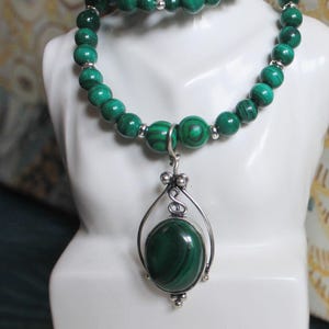 Peut inclure: Un ensemble collier et bracelet en perles de malachite verte. Le collier présente un pendentif en argent avec une grosse pierre ovale en malachite. Les perles sont d'un vert profond, et le collier est présenté sur un buste blanc.