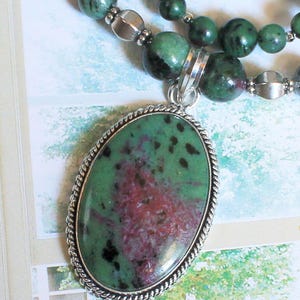 Puede incluir: Un collar con un gran colgante ovalado de piedra verde y magenta engastado en un marco de metal plateado trenzado. El collar está compuesto por cuentas redondas verdes y magenta intercaladas con pequeñas cuentas espaciadoras de metal plateado.