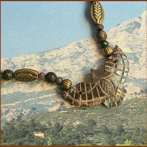 Peut inclure: Un pendentif en pierre marron et verte en forme d'oiseau avec des détails complexes. Le pendentif fait partie d'un collier avec des perles brunes, vertes et rouges et des accents de feuilles dorées.