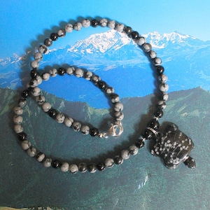 Può includere: Collana di perline in ossidiana fiocco di neve grigia e nera, con chiusura argentata. La collana presenta un ciondolo a forma di tartaruga in ossidiana fiocco di neve. Lo sfondo mostra un paesaggio montano.