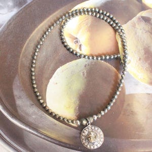 Peut inclure: Un collier composé d'un rang de petites perles facettées gris métallisé. Un pendentif circulaire est suspendu au centre, présentant une pierre centrale aux tons dorés texturés entourée d'un bord en métal argenté orné de petits points.