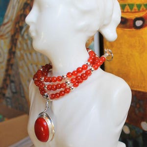 Peut inclure: Un collier à plusieurs rangs composé de petites perles rondes rouge vif alternées avec des sphères métalliques argentées polies. Le collier comprend un grand pendentif cabochon rouge de forme ovale serti dans une monture en métal argenté.