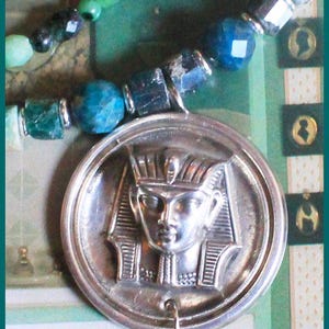 Peut inclure: Un pendentif en argent représentant un relief détaillé de la tête d'un pharaon avec un couvre-chef. Le pendentif est attaché à un collier avec des perles turquoise et noires.