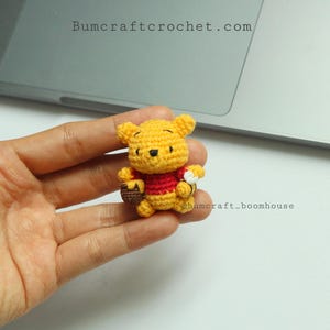 Pode incluir: Pequena figura de crochê do Ursinho Pooh, amarela e vermelha. O urso está segurando uma pequena cesta e uma flor. A figura é feita à mão e tem os textos "bumcraftcrochet.com" e "@bumcraft_boomhouse".