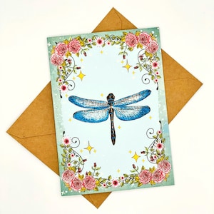 May include: A blue dragonfly with black markings is surrounded by a floral border of pink and white flowers on a light blue background. The border is made of vines and leaves. The background has a subtle watercolor effect.