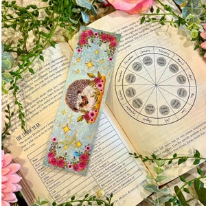 May include: A blue and white bookmark with a hedgehog and flowers. The bookmark has a list of trees and their corresponding lunar months. The bookmark is on top of a book with a wheel showing the different moon phases throughout the year.