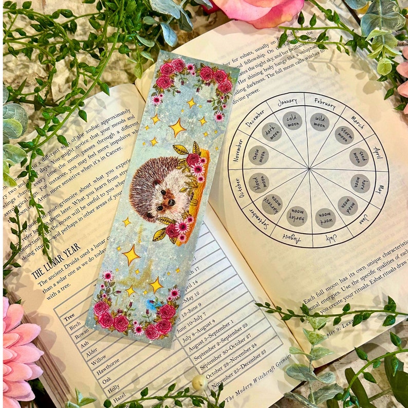 May include: A blue and white bookmark with a hedgehog and flowers. The bookmark has a list of trees and their corresponding lunar months. The bookmark is on top of a book with a wheel showing the different moon phases throughout the year.
