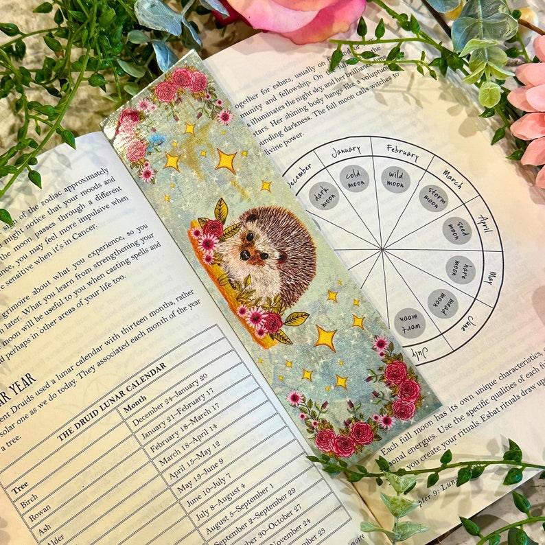 May include: A blue and gold bookmark with a hedgehog illustration, pink flowers, and gold stars. The bookmark features a circular chart with the months of the year and moon phases. The text on the bookmark reads "The Druid Lunar Calendar".