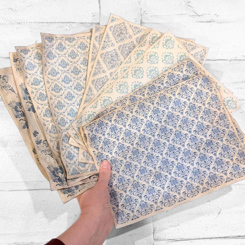 May include: A hand holds a fan of vintage-style paper sheets. Each sheet features a blue floral damask pattern on a cream background. The papers vary slightly in color and pattern, creating a layered effect. The papers are rectangular and appear aged.