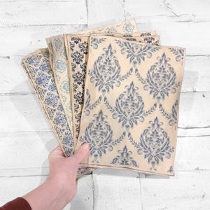 May include: A hand holds several sheets of patterned paper. The papers feature a repeating blue floral design on a cream background. The patterns vary slightly in color and style, with some having a more vintage or distressed look.