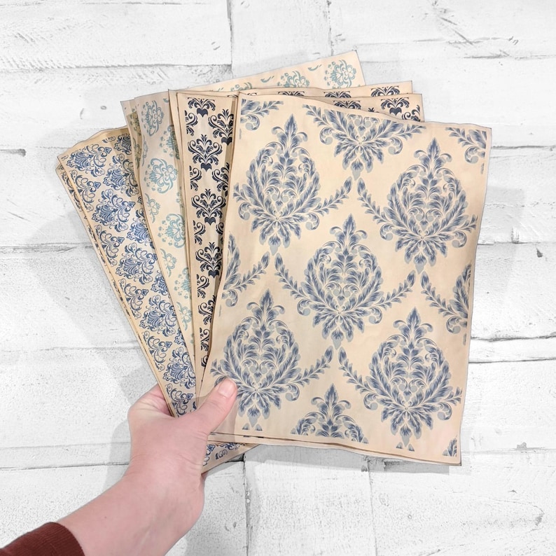 May include: A hand holds several sheets of patterned paper. The papers feature a repeating blue floral design on a cream background. The patterns vary slightly in color and style, with some having a more vintage or distressed look.