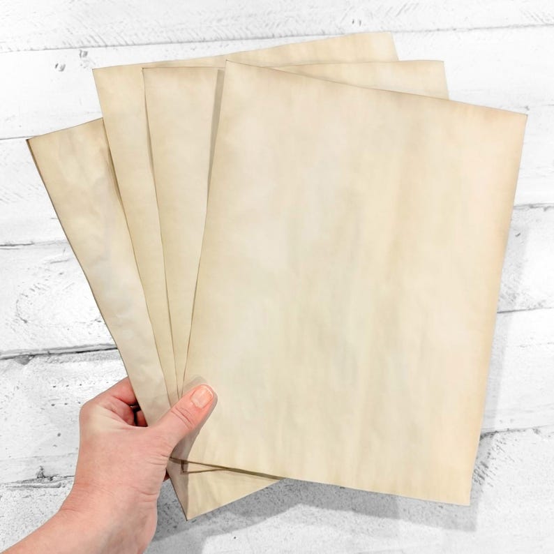 May include: A hand holding a stack of aged, cream-colored paper sheets. The paper has a vintage look with subtle brown staining around the edges, suggesting an antique or weathered appearance. The background is a white wooden surface.