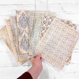 May include: A collection of patterned paper sheets in various designs and shades of blue and cream. The papers feature floral and damask patterns, with some sheets showcasing a more intricate design. The papers are held in a hand.