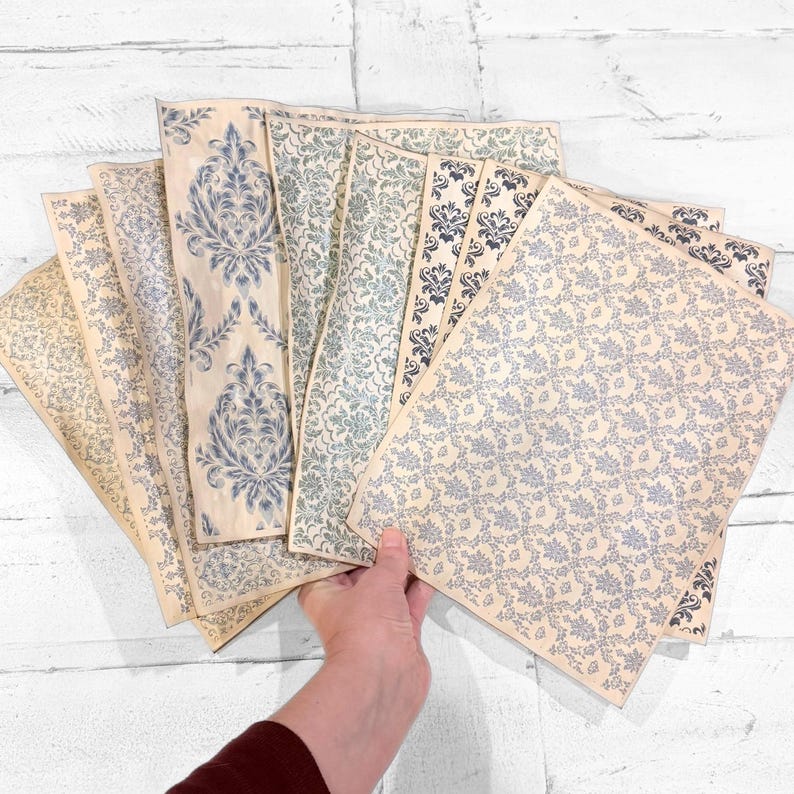 May include: A collection of patterned paper sheets in various designs and shades of blue and cream. The papers feature floral and damask patterns, with some sheets showcasing a more intricate design. The papers are held in a hand.