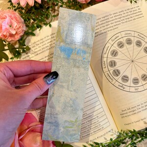 May include: A vintage-style bookmark with a watercolor design of a blue and green landscape. The bookmark is made of paper and has a slightly textured surface.