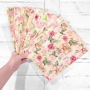 May include: A collection of floral-patterned papers featuring pink roses and green leaves on a cream background. The papers are held in a fan shape, showcasing the variety of designs. The overall aesthetic is vintage and romantic.