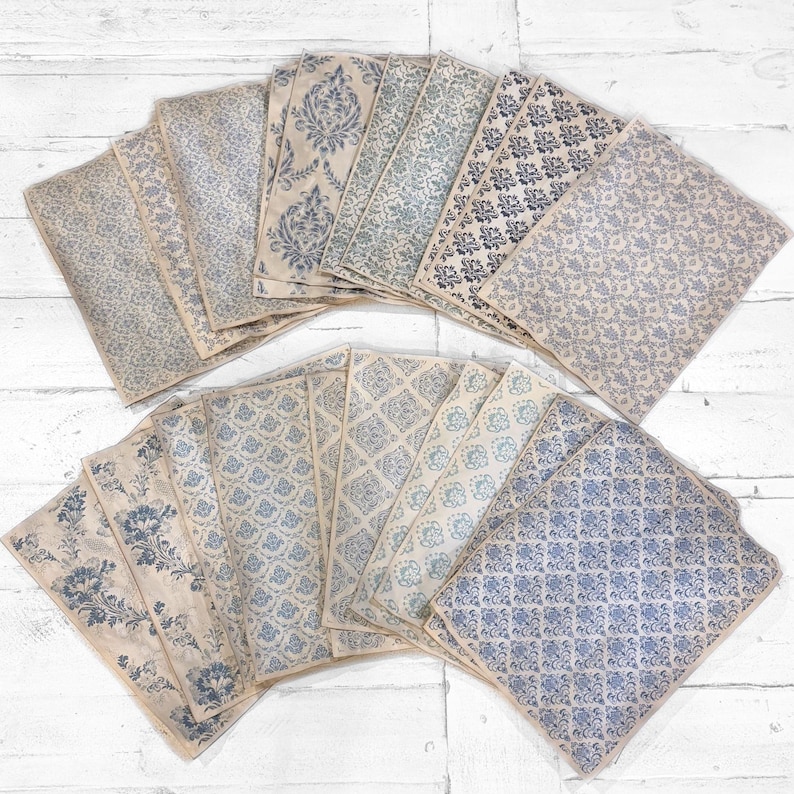 May include: A collection of vintage-style paper sheets with various blue floral and geometric patterns. The papers have a distressed, aged appearance, with a beige background and blue designs. The sheets are arranged in a fan shape.