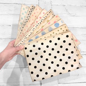 May include: A collection of patterned paper sheets fanned out, featuring various polka dot designs. The sheets have a vintage, aged appearance with black, blue, pink, and gold dots on a cream background. The paper is held in a hand against a white wooden backdrop.