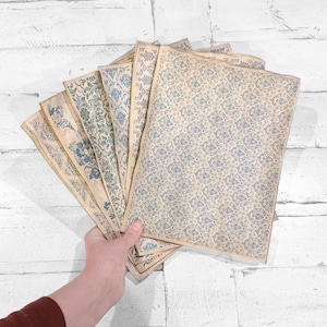 May include: A collection of vintage-style paper sheets with blue floral patterns. The sheets are arranged in a fan shape, showcasing various designs. The paper has a slightly aged, off-white color, and the floral patterns vary in style.