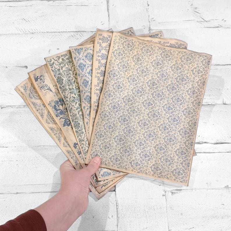 May include: A collection of vintage-style paper sheets with blue floral patterns. The sheets are arranged in a fan shape, showcasing various designs. The paper has a slightly aged, off-white color, and the floral patterns vary in style.