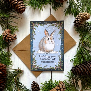 May include: A greeting card with a blue background and a white rabbit illustration. The card has a green border with holly and berries. The text on the card reads "Wishing you a season of coziness!"