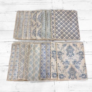 May include: A collection of vintage-style paper sheets with various blue and cream-colored patterns. The designs include floral, damask, and geometric motifs, arranged in rows on a white wooden surface.