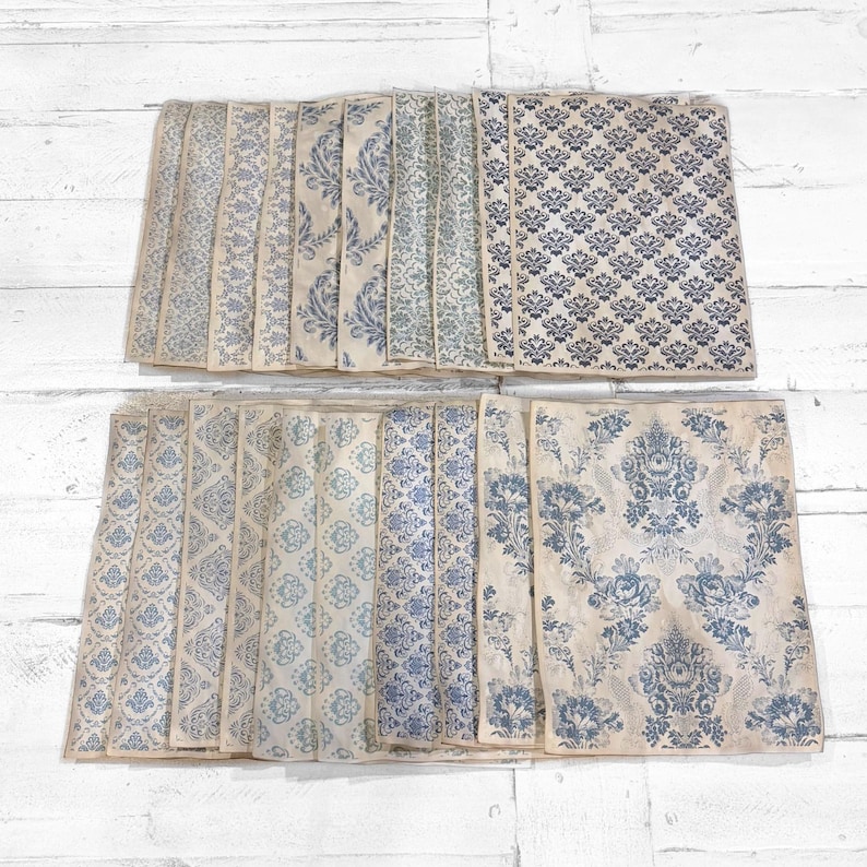 May include: A collection of vintage-style paper sheets with various blue and cream-colored patterns. The designs include floral, damask, and geometric motifs, arranged in rows on a white wooden surface.